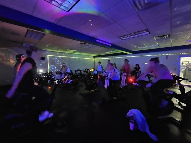 Members riding stationary bikes during a high-energy indoor spin class at the Sinking Spring YMCA, with low lighting and LED accents.