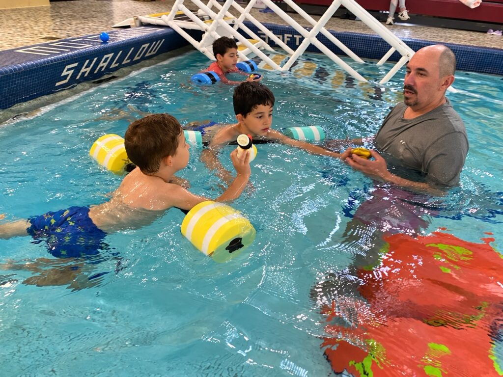 Swim instructor works with three children using flotation devices during a lesson in the shallow end of an indoor pool.
