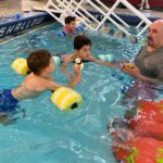 Swim instructor works with three children using flotation devices during a lesson in the shallow end of an indoor pool.